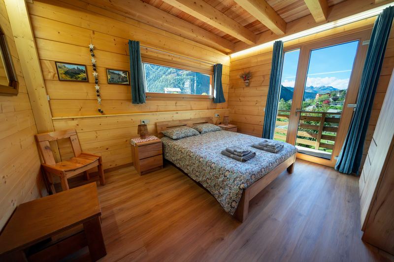 Camera Tirlo di giorno: letto matrimoniale sotto la finestra a nastro, pareti e soffitto in legno, portafinestra aperta sul terrazzo con le montagne